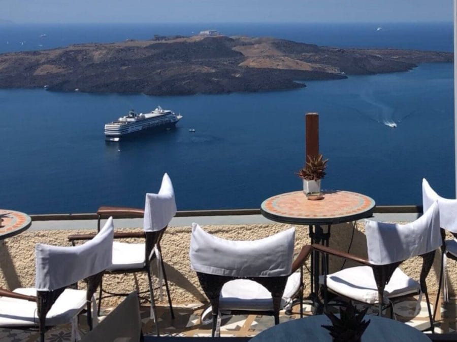 A restaurant in Fira, Santorini, with outdoor seating and the view of Nea Kammeni island and a cruise ship sailing in the Aegean Sea