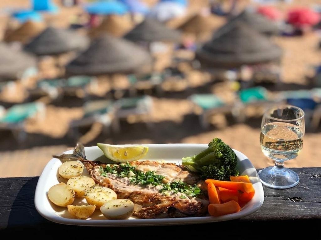 A white plate with some fish, potatoes, broccoli, carrots and lime, and a glass of white wine and umbrellas, with chairs on Praia da Coelha in the background
