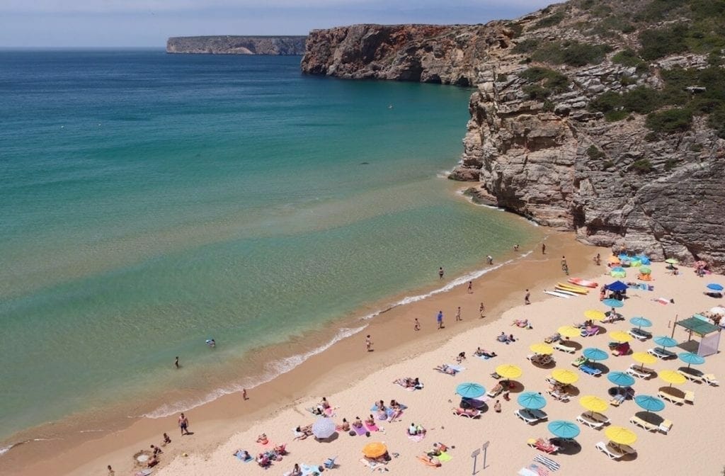 Umbrellas of different colours and people sunbathing on Praia do Beliche, Sagres, that is bordered by colossal limestone cliffs and has crystalline green water 