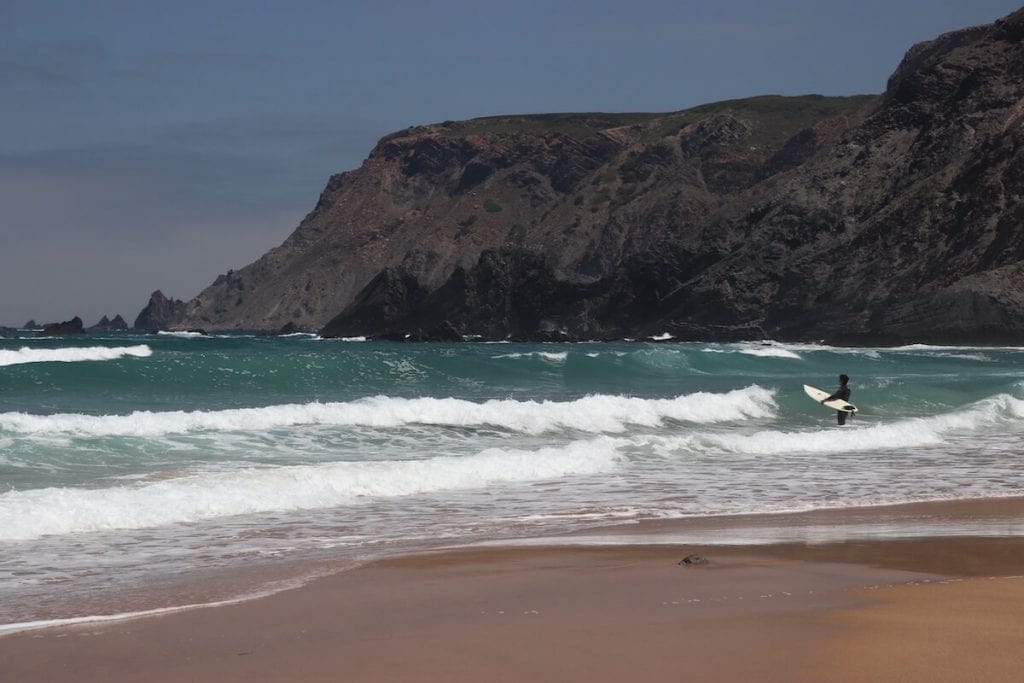A man with a surfboard getting into the water on a beach in Sagres, Portugal
