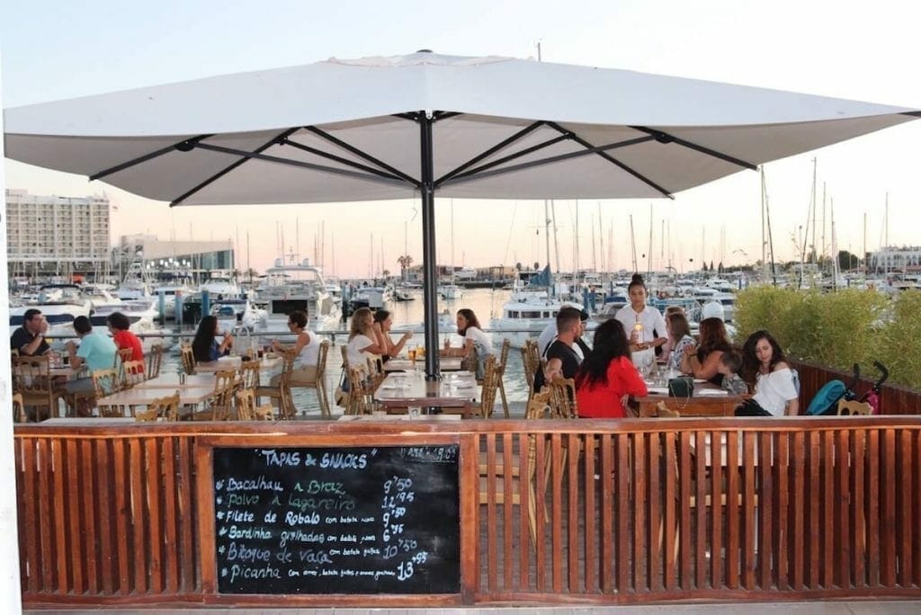 People having drinks and talking in a bar with outdoor tables and chairs and a large white umbrella located at Vilamoura Marina, with the marina and some boats in the background