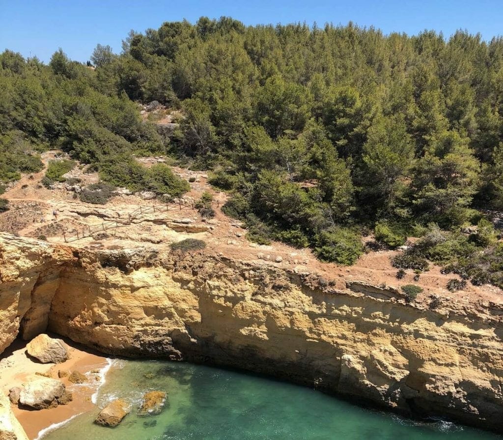 Part of the Seven Hanging Valleys Trail, Algarve, with a small beach with crystal-clear water surrounded by orange limestone cliffs some local vegetation covering the cliffs
