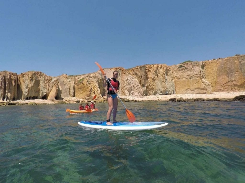 A woman doing Stand-up paddle boarding and three people on an orange kayak on Praia dos Arrifes, Albufeira, and some orange cliffs in the background