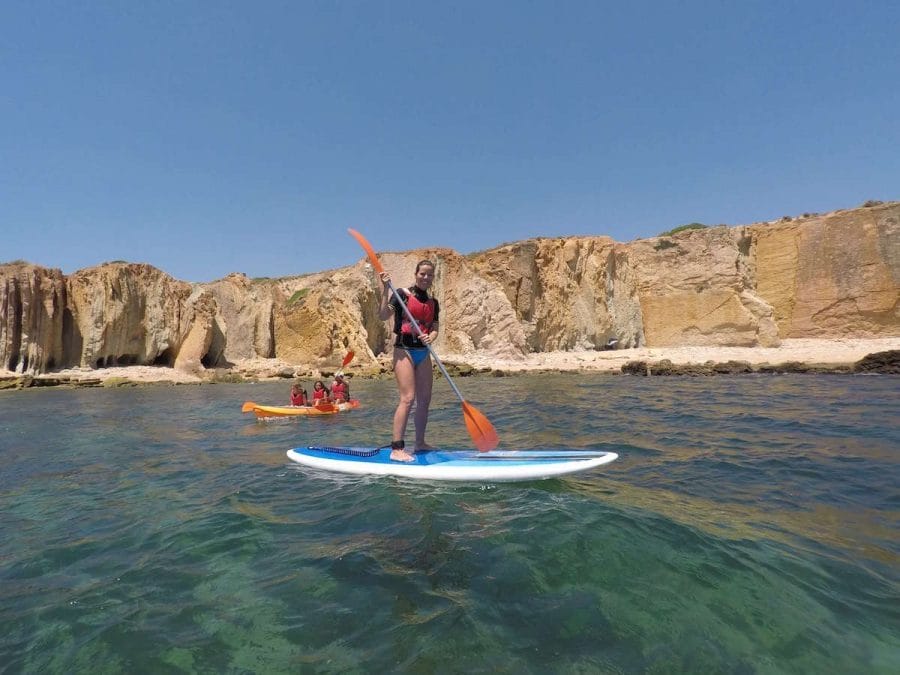 A woman wearing a red lifejacket and blue bikini on a Stand up paddle board and three people on a kayak sailing along the Algarve Coast