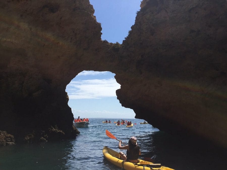 A woman kayaking along the Algarve coast and passing through a sea arch towards a boat with some people wearing red lifejackets and other kayaks