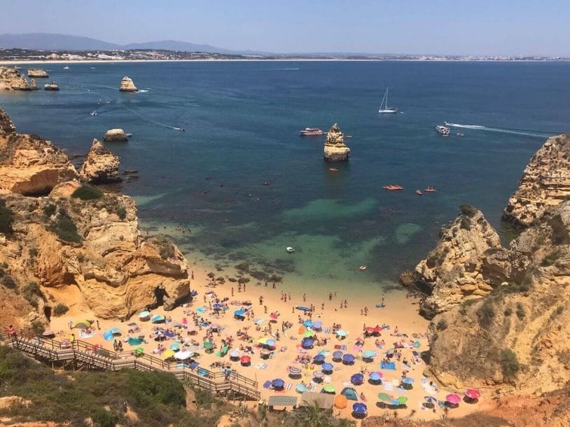 Praia do Camilo, Lagos, Portugal, with many umbrellas with different colours, people walking on the beach that's surrounded by massive yellow-orangish limestone cliffs and a wooden stairway that gives access to the beach