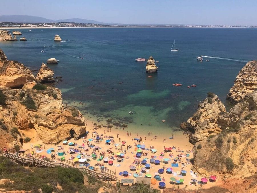 Praia do Camilo, Lagos, enclosed by orange-yellowish limestone cliffs, with many umbrellas of different colours on the beach, a wooden staircase and a few boats and kayaks sailing in the crystal-clear water