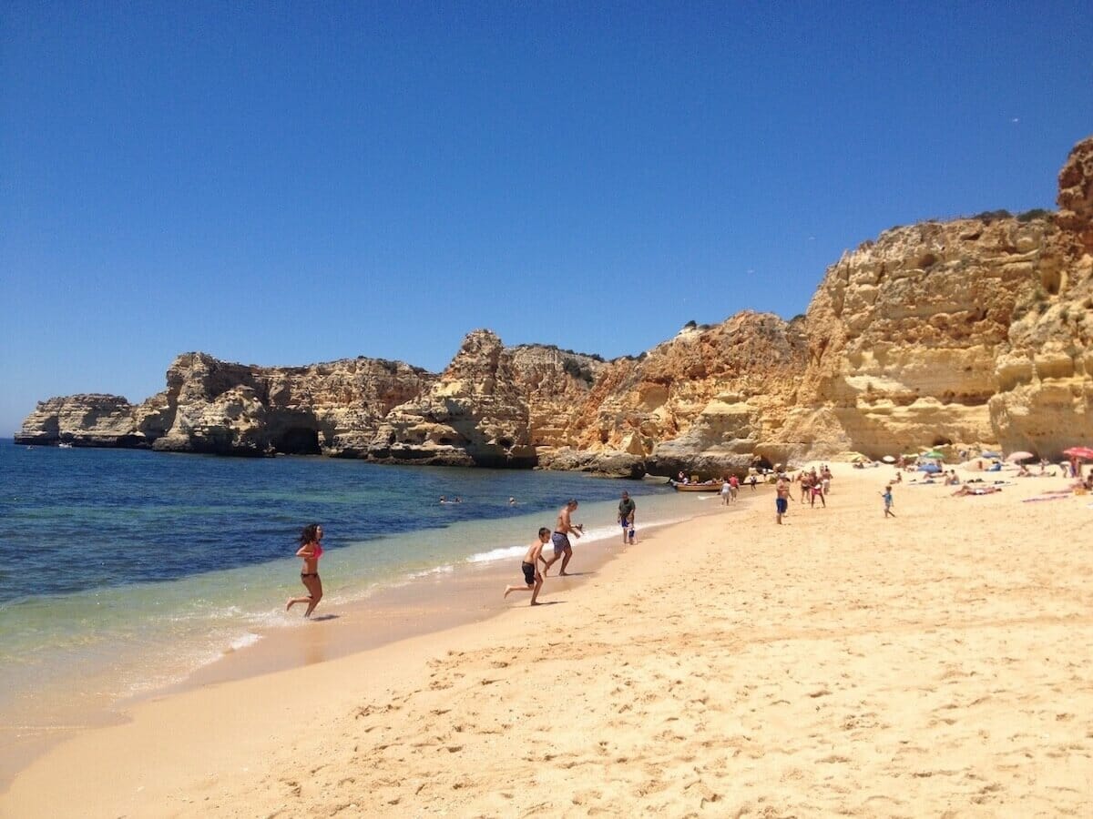 People walking on the amber sand soft sand of Praia da Marinha, that's bordered by massive yellow-orangish cliffs and has crystalline blue water.