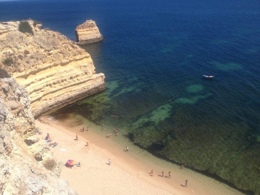 The view of Praia da Marinha, Algarve, Portugal, from the pathway with people walking on the beach and swimming on its crystalline blue water, and yellow limestone cliffs.