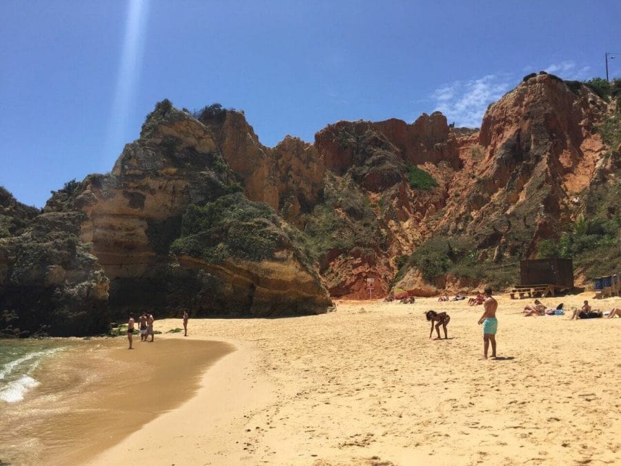 People walking on the amber sand of Praia do Camilo, Lagos, Portugal, during the low season and some red limestone cliffs in the background.