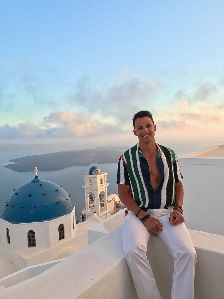 30 Amazing Things to Do in Santorini, Greece 23 Pericles Rosa wearing a striped shirt and white trouser sitting on a white balcony in the village of Imerovigli, Santorini, and ti the background the Anastasi Church, Nea Kammeni island and the Aegean Sea