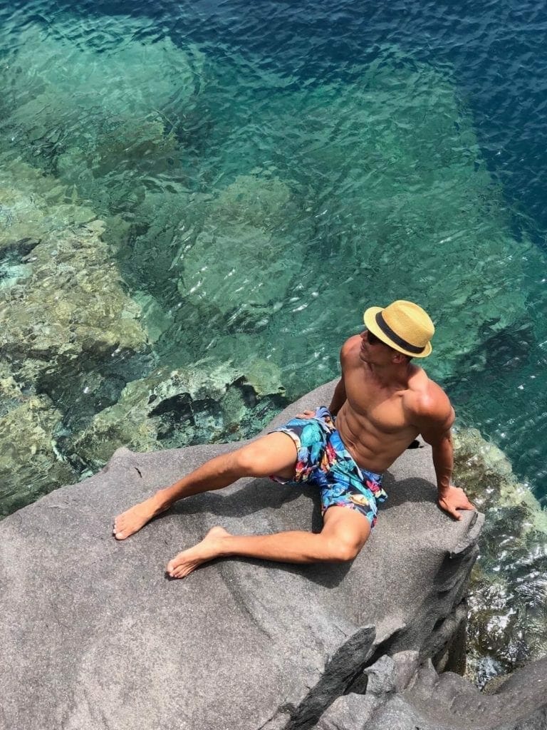 A man wearing a hat, sunglasses and colourful short sitting on a rock at Amoudi Bay, Santorini, Greece