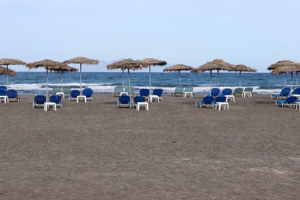 A stretch of black sand with umbrellas and beaches chairs on Monolithos Beach, Santorini, Greece