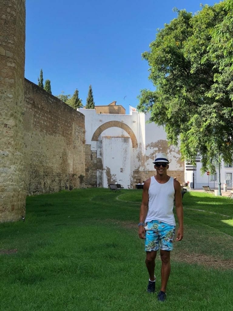um homem usando um chapéu branco, uma regata branca e um short colorido andando perto das antigas muralhas da cidade velha de Faro