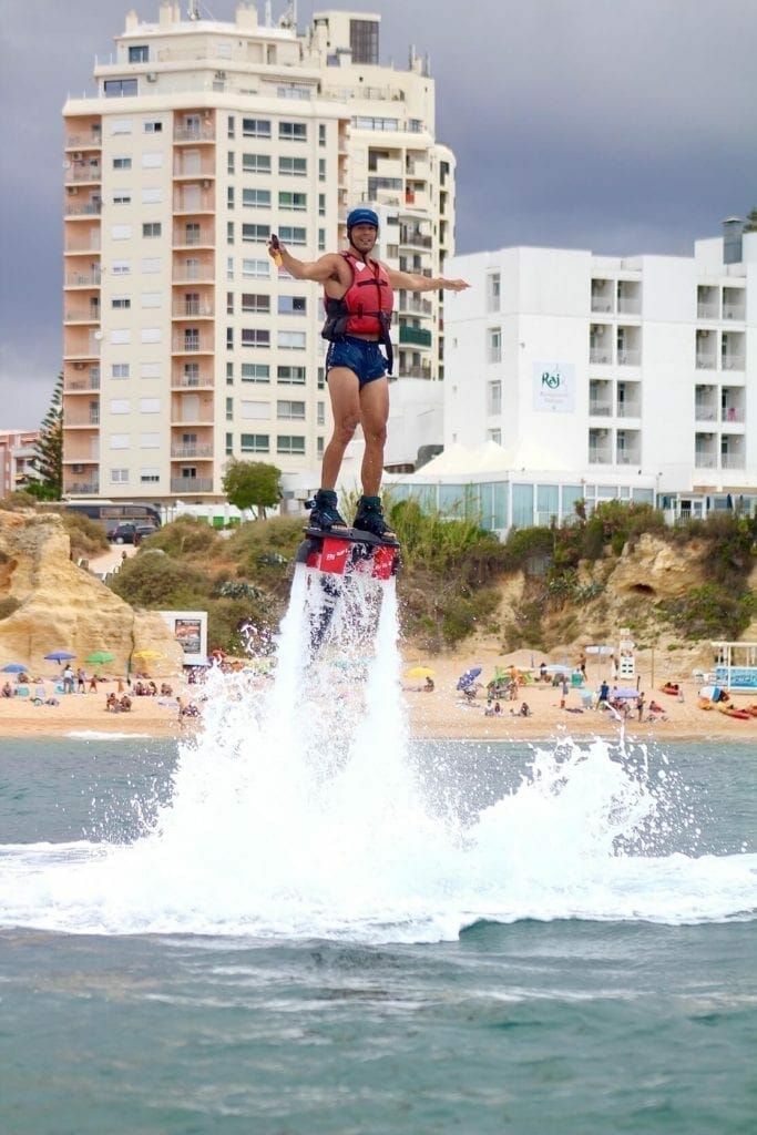 A man wearing a blue short, red lifejacket and blue helmet flying above the Algarve coast on a flyboard in Praia de Armacão de Pêra