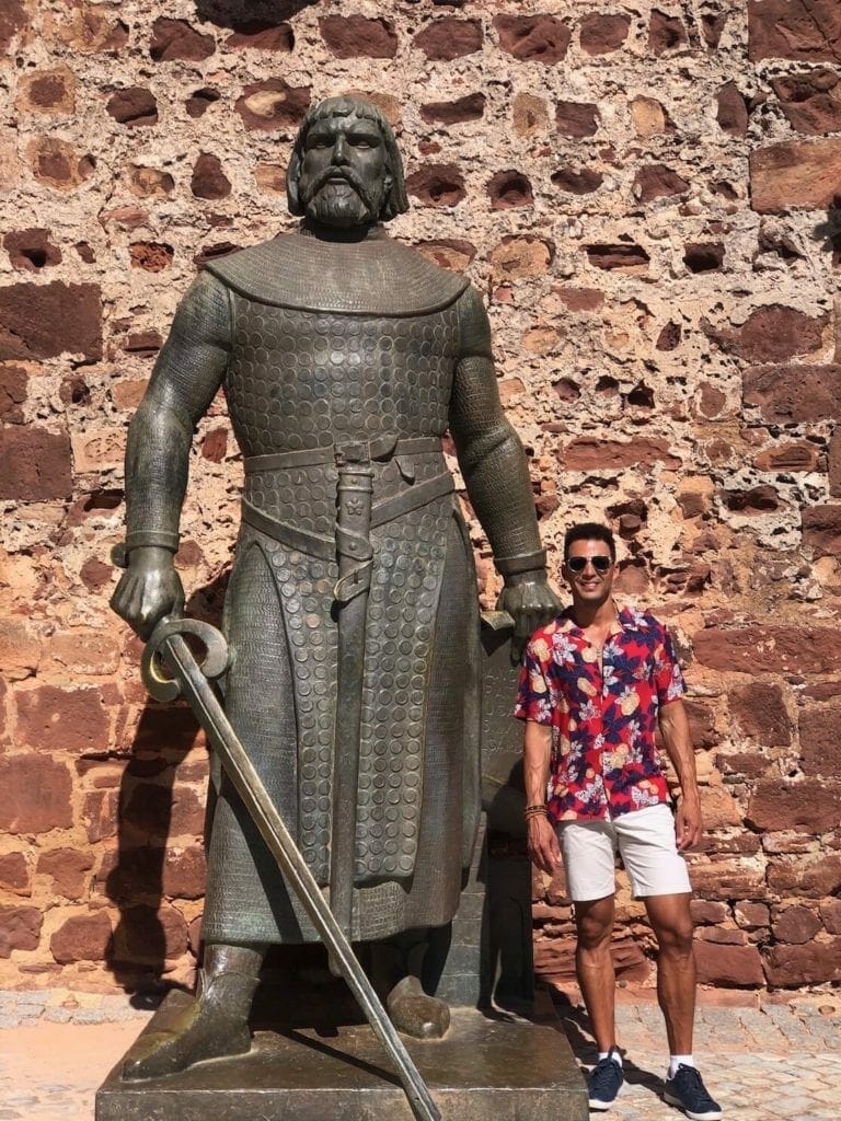 Pericles Rosa wearing a beige short and floral red shirt with a huge statue of King D. Sancho I at the entrance of Silves Castle, Portugal