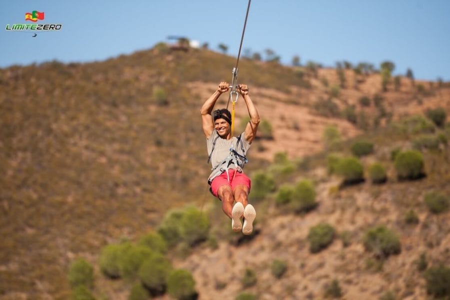 Pericles Rosa wearing a brown t-shirt and red short ziplining from Spain to Portugal with a hill covered with sparsed trees in the background