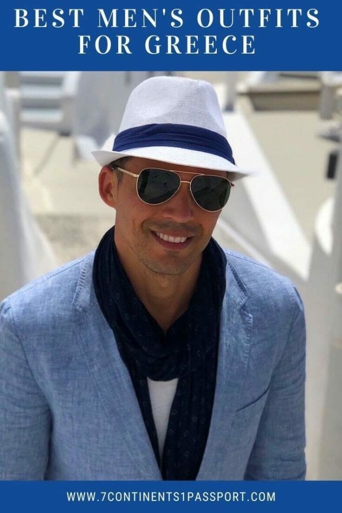 A man wearing a white hat, sunglasses, marine-blue scarf, white t-shirt and light-blue blazer on an alley in Imerovigli, Santorini, with some white-washed houses in the background