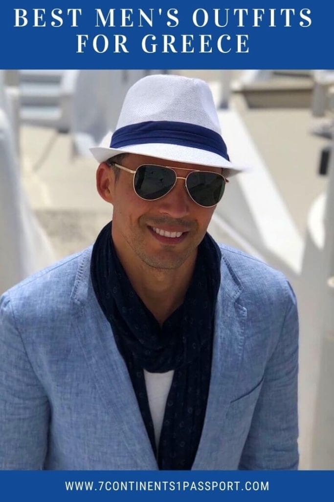 What to Wear in Santorini – Best Men's Outfits for Greece 11 A man wearing a white hat, sunglasses, marine-blue scarf, white t-shirt and light-blue blazer on an alley in Imerovigli, Santorini, with some white-washed houses in the background