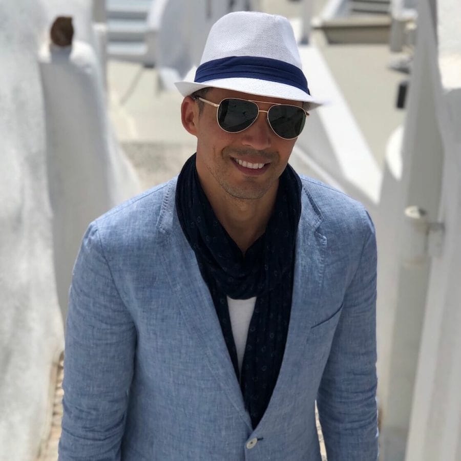Pericles Rosa wearing a white hat, sunglasses, marine-blue scarf, white t-shirt and light-blue blazer on an alley in Imerovigli, Santorini, with some white-washed houses in the background