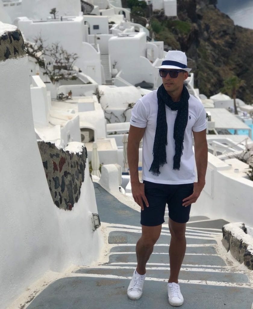 Pericles Rosa wearing one of his Santorini outfits, composed of a white hat, sunglasses, a marine blue scarf, white polo, marine blue shorts and white trainers in an alley in Oia, Santorini, Greece. 