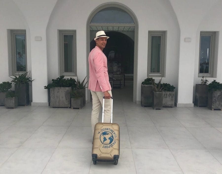 Pericles Rosa wearing a white hat, salmon blazer, beige trouser carrying a hand-luggage in front of the entrance of a hotel with white walls in Santorini, Greece