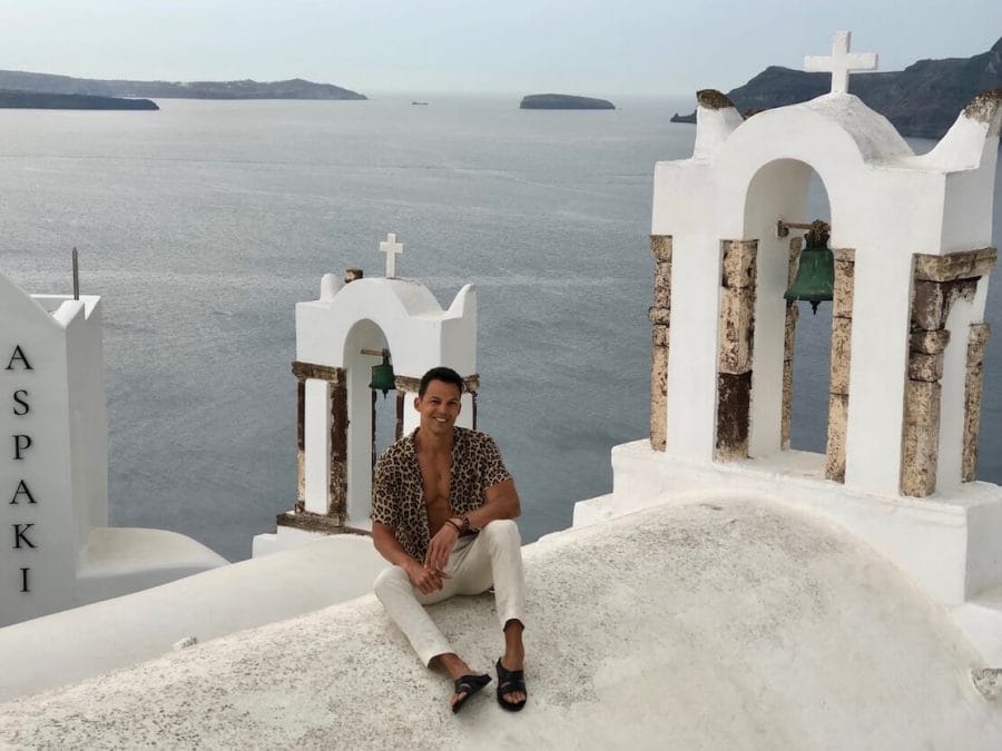 Pericles Rosa wearing a leopard print shirt, beige linen trousers and sandals sitting on the top of a church in Oia, Santorini, with two bell towers and the Aegean Se behind him