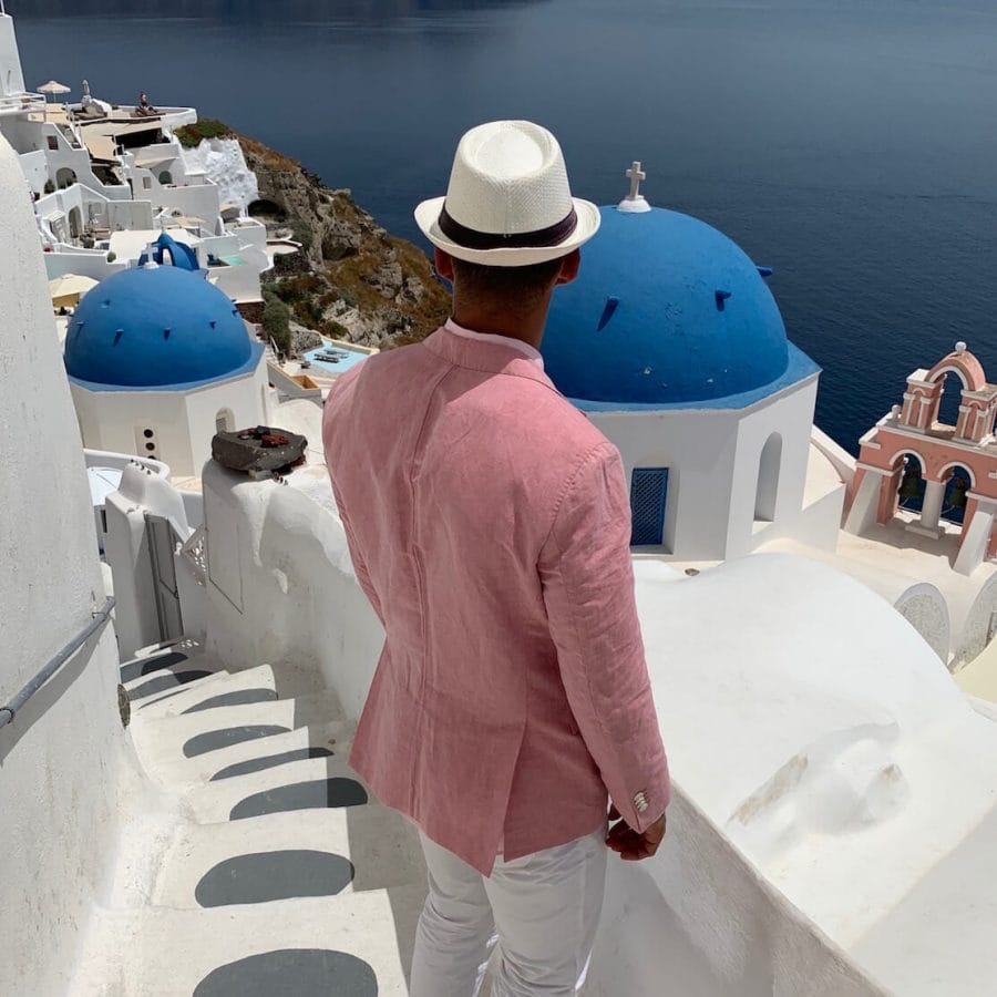 Pericles Rosa wearing one of his Greek outfits for males. In this composition, he's wearing a white hat, salmon blazer and white trousers on one stairway in Oia, Santorini, with some whitewashed houses and blue-domed churches. 