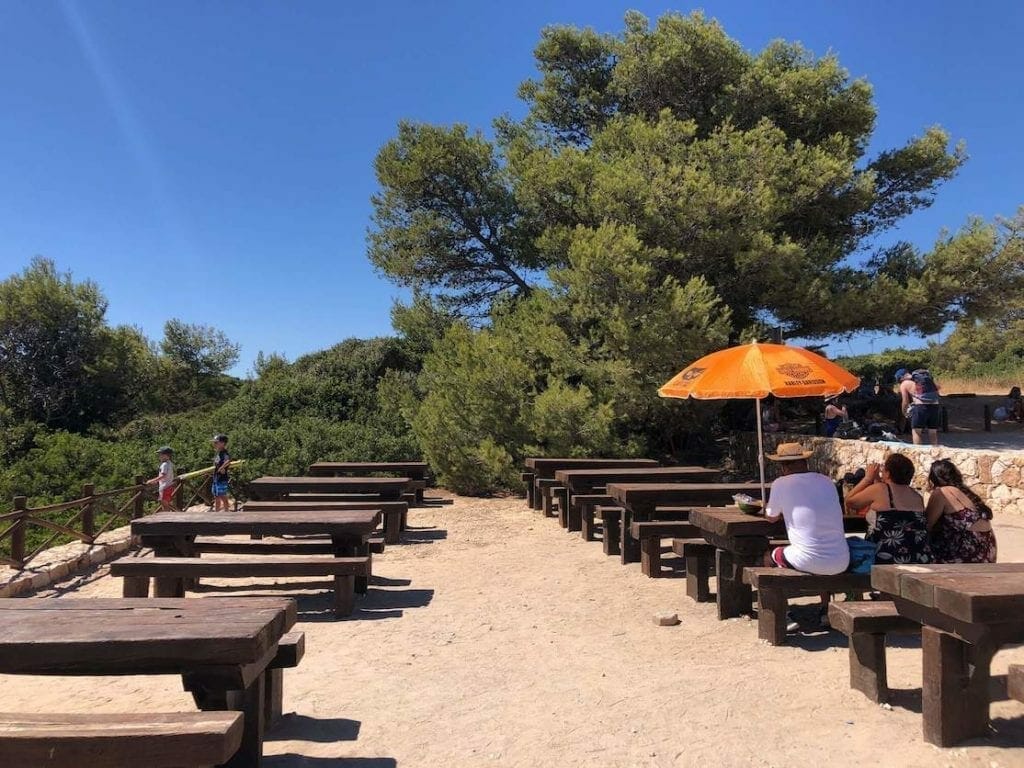 Uma área de piquenique no final da Trilha dos Sete Vales Suspensos no Algarve, Portugal, com mesas e bancos de madeira, algumas árvores e pessoas em uma das mesas com um sombreiro laranja bebendo e comendo