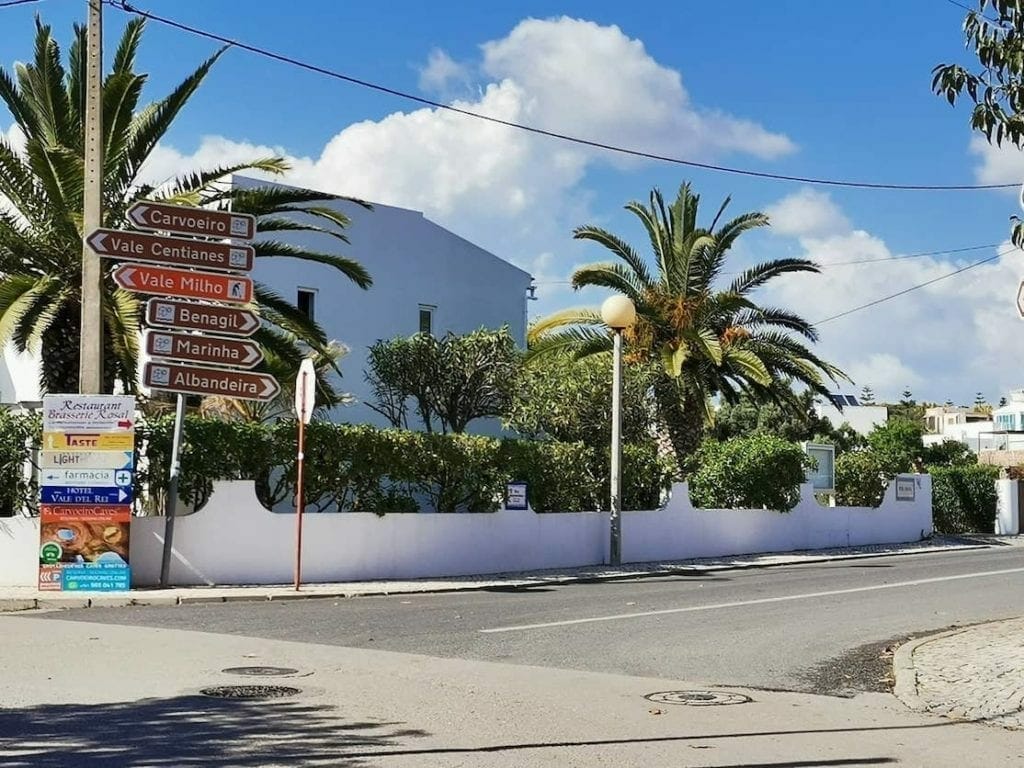 Algumas placas na esquina de uma rua mostrando como chegar à Praia da Marinha, Carvoeiro, Benagil e Albandeira, uma casa branca e árvores ao fundo