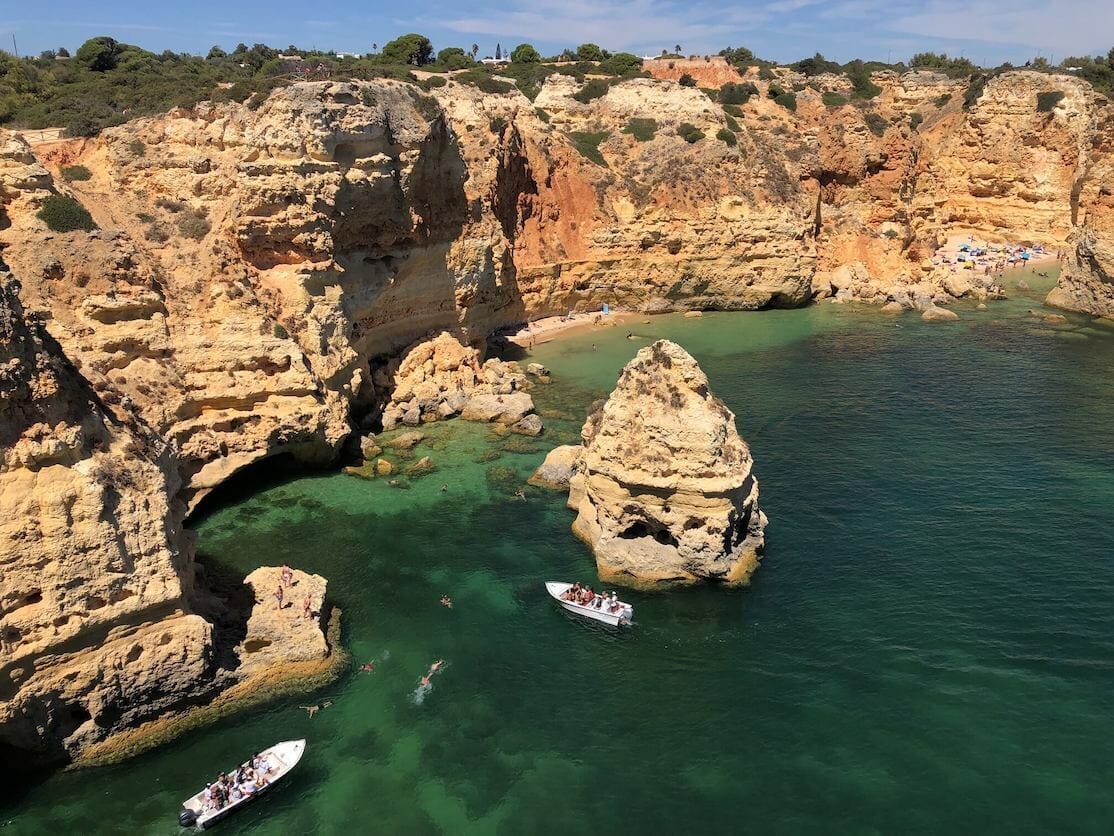 La extraordinaria playa de Marinha, una de las playas más bellas del Algarve, Portugal, con gente nadando y dos barcos en sus aguas cristalinas azul bordeadas por acantilados anaranjados-amarillentos