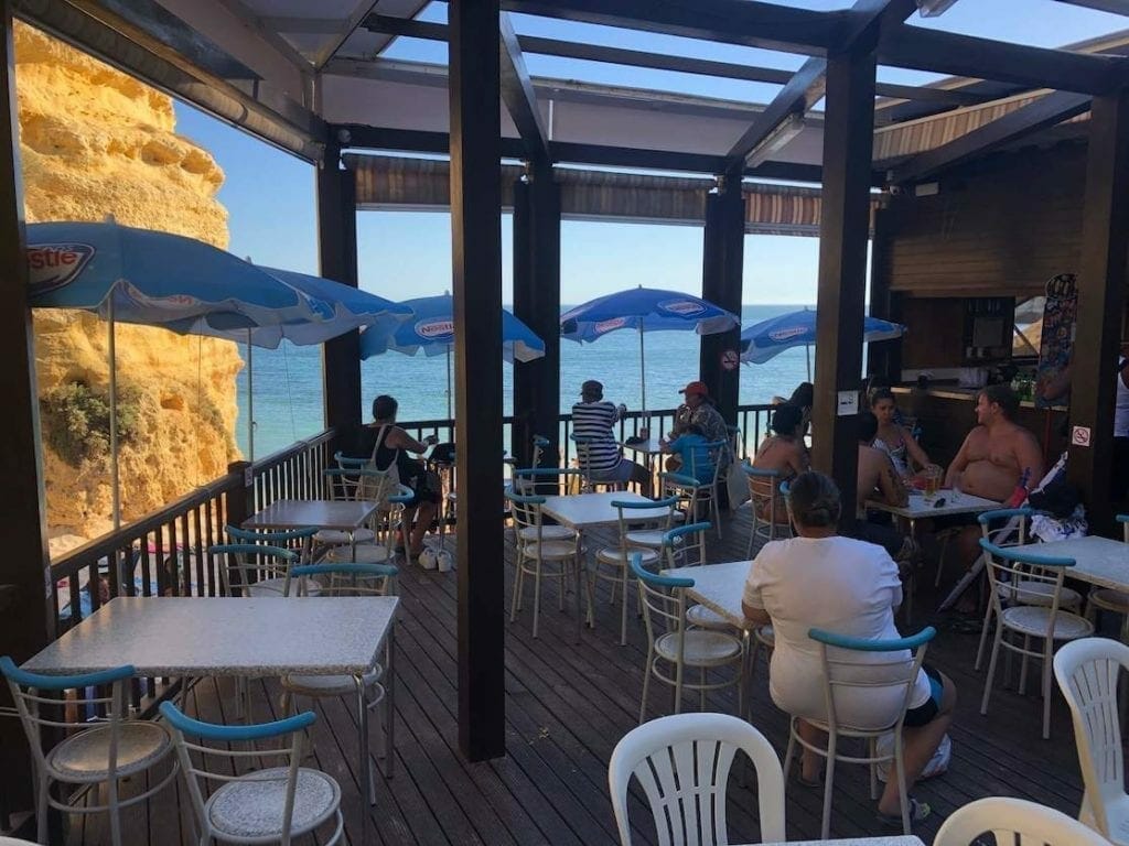 Restaurante à beira-mar da Praia da Marinha com algumas mesas, cadeiras, guarda-chuvas e pessoas curtindo a vista e conversando