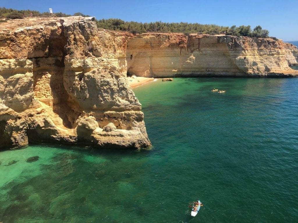 Praia do Corredoura, no Algarve, Portugal, rodeada por falésias amarelas cobertas de vegetação baixa, e três caiaques a navegar na sua água cristalina esmeralda