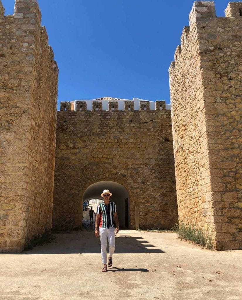 Pericles Rosa wearing a beige trouser, striped shirt and a hat walking in Old Town Lagos, Portugal, and the ancient city walls behind him