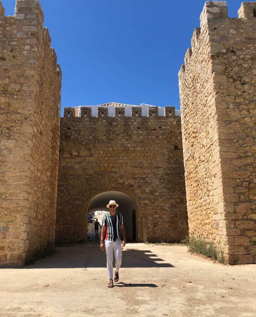 18 Best Things to Do in Lagos, Portugal 8 Pericles Rosa wearing a beige trouser, striped shirt and a hat walking in Old Town Lagos, Portugal, and the ancient city walls behind him