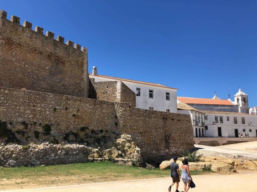 A man and a woman walking near the rocky walls of Lagos castle towards a white church in downtown, Lagos, Portugal