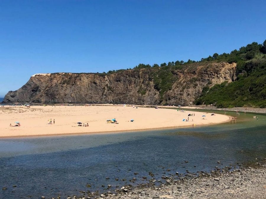 Praia de Odeceixe, Portugal, e a sua extensa faixa de areia de forma arredondada, um rio de águas cristalinas margeado por uma falésia negra coberta com vegetação que circunda à praia