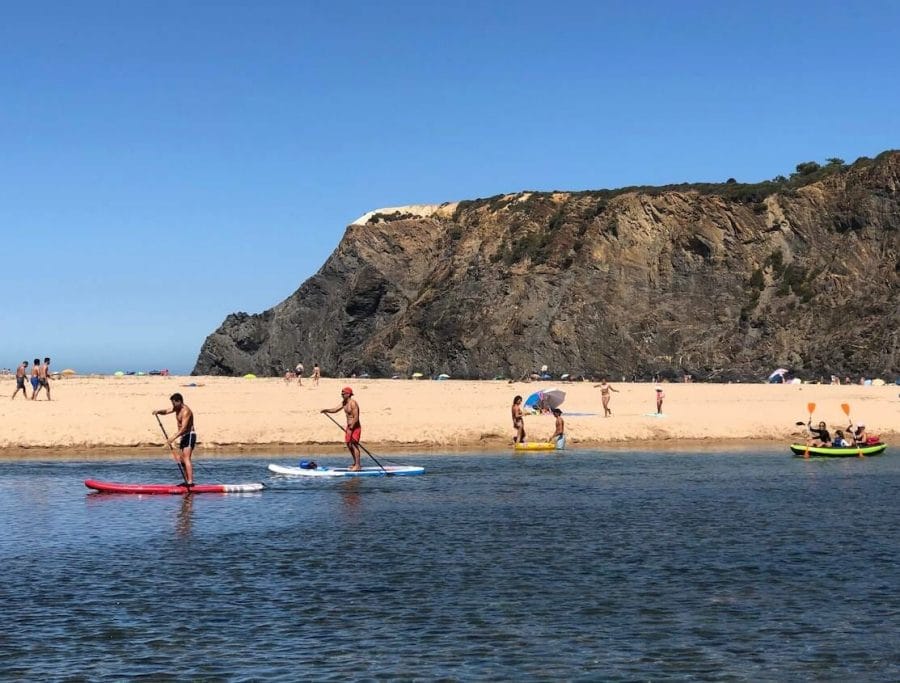 People on SUP and on an inflatable paddling along a river at Praia de Odeceixe, and others walking on the sandbank and a massive cliff in the background