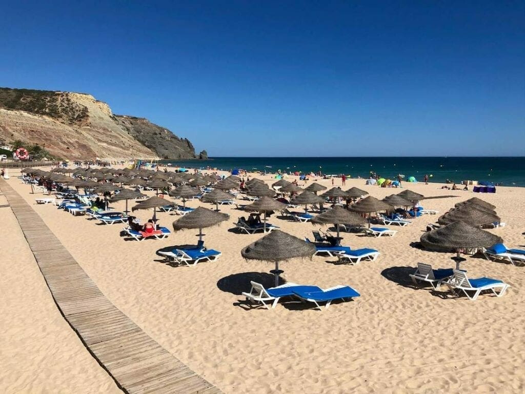 A wooden boardwalk, umbrellas and blue lounge chairs on Praia da Luz, that has soft sand, crystal-clear water and is bordered by an imposing basalt cliff.