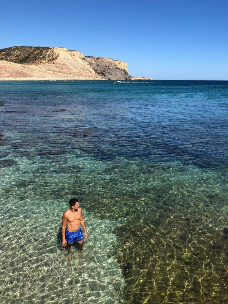Péricles Rosa vestindo um short azul na água cristalina da Praia da Luz, Portugal, e ao fundo a falésia cinza que margeia a praia