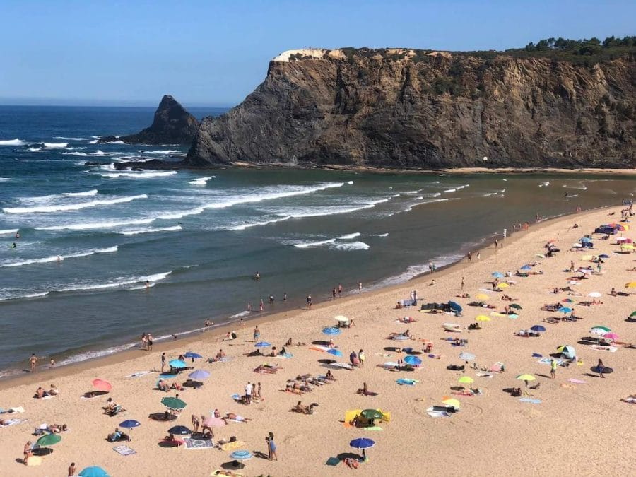 People sunbathing on towels and underneath umbrellas of different colours on Praia de Odeceixe, Portugal, the sea and a black cliff topped with vegetation in the background
