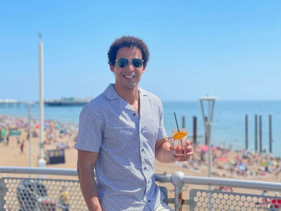 Péricles Rosa usando óculos escuros e shorts brancos pontilhados com pontos azuis tomando uma bebida no Brighton Beach Club e na praia atrás dele