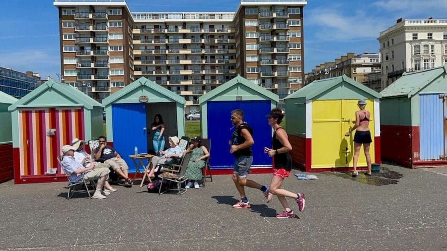 um casal correndo e algumas pessoas sentadas na frente de algumas cabanas de praia coloridas em Hove, Brighton, Inglaterra