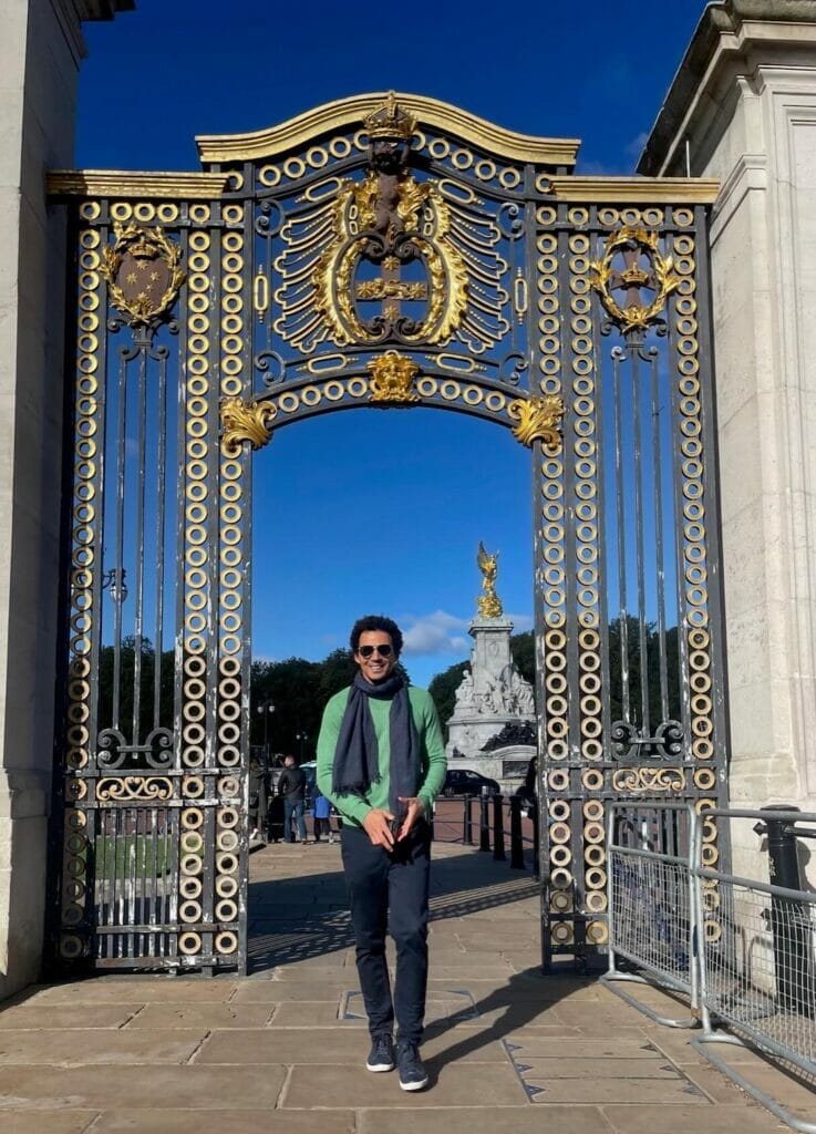 Péricles Rosa usando óculos de sol, um suéter verde, cachecol azul, calças azuis marinhas e tênis passando por um portão do St. James's Park, Londres, e o Queen Victoria Memorial ao fundo
