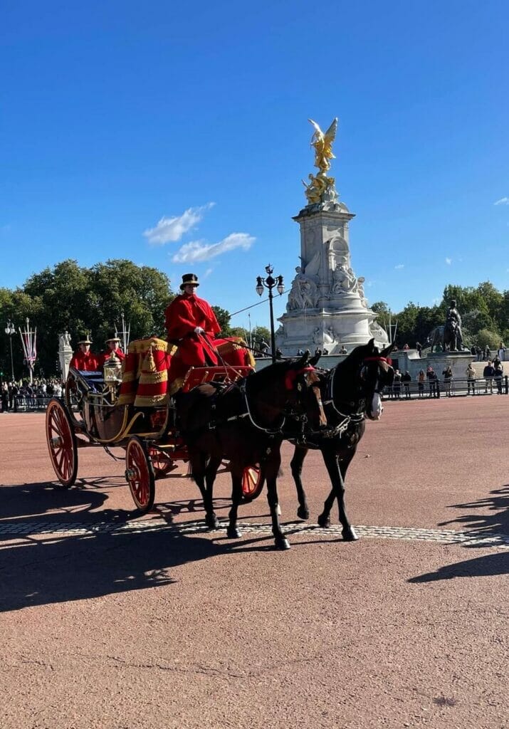 Dois cavalos, uma carruagem e três homens usando capa vermelha e chapéu preto em frente ao Queens Victoria Memorial, londres, trazendo convidados para visitar o Palácio de Buckingham