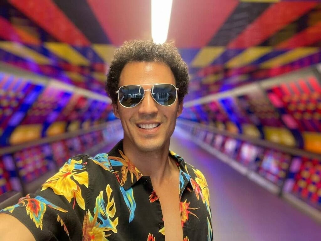 Pericles Rosa wearing sunglasses and a colourful shirt taking a selfie at Adams Plaza Bridge in Canary Wharf, London, which was painted with vivid colours by Camille Walala