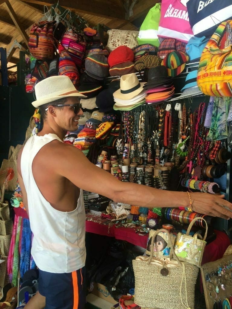 A man wearing a hat, sunglasses, a white tank top and blu shorts shopping at Straw Market, Downtown Nassau, the Bahamas