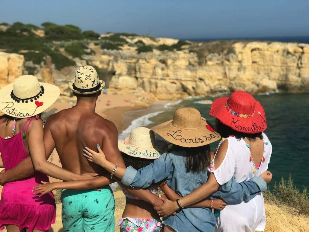 Cinco pessoas com chapéus personalizados de costas abraçadas admirando a vista de um mirante na Praia da Coelha em Albufeira, Portugal