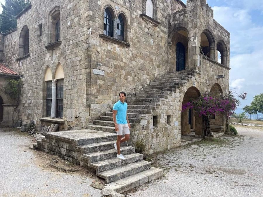 Pericles Rosa wearing a blue polo, beige shorts and white trainers on the steps og The Monastery of Filerimos, Ancient City of Ialysos, Greece