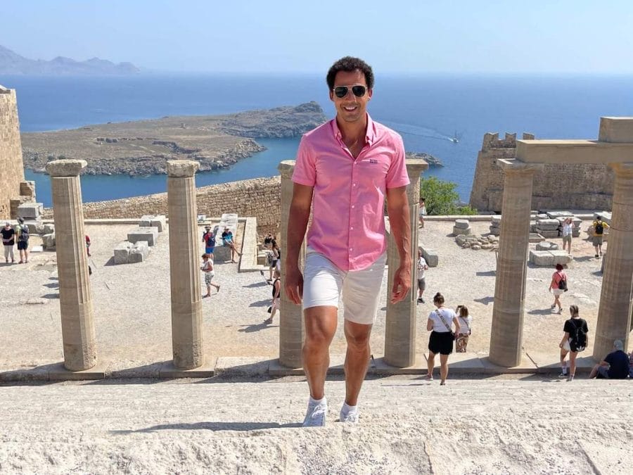 17 Pictures to Prove That Rhodes Should Be Your Next Greek Destination 9 Pericles Rosa wearing sunglasses, salmon shirt, beige shorts and white trainers, posing for a picture ar Lindos Acropolis, Rhodes, Greece